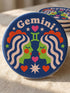 A close-up photograph of a round, glossy ceramic coaster featuring a bold retro pop art design. The background is a deep royal blue. In the center are two symmetrical profile faces of twins looking at each other, featuring bright green skin, closed eyes, and red cheeks. They have wavy white and pale pink hair with red accents. The word Gemini is arched at the top in a retro white font. The design is decorated with a green Gemini symbol, pink and red hearts, and white starbursts.
