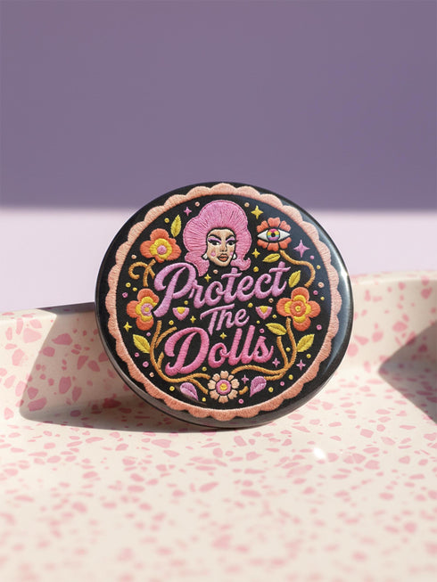 A circular black pinback button featuring the slogan Protect The Dolls in retro script. The design utilizes a high-fidelity trompe-l'œil print style that mimics the texture of raised thread embroidery with satin-stitch detailing. The artwork features vibrant hot pink, rust orange, and golden yellow motifs including drag queens with bouffant hair, 70s style daisies, lightning bolts, and a crescent moon with a face. The surface is smooth and glossy, creating a faux-textured illusion.