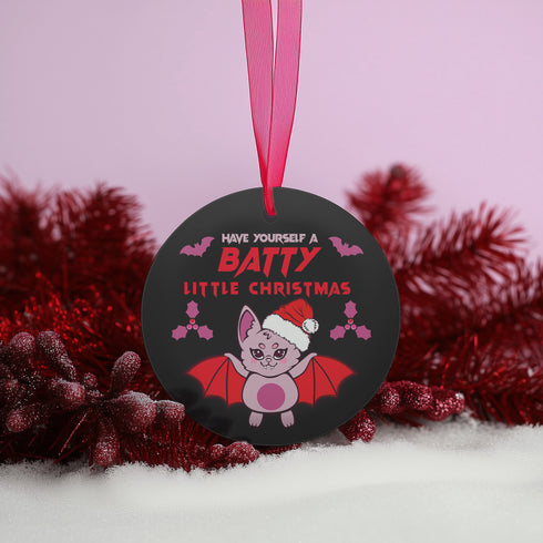 Round black metal Christmas ornament featuring cute pink bat with red wings wearing Santa hat, surrounded by pink bats and holly. Text reads "Have Yourself A Batty Little Christmas"