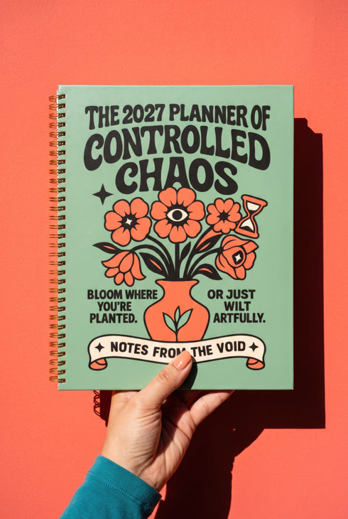 A sage green hardcover spiral planner for 2027-2028 with the title "The 2027 Planner of Controlled Chaos" in a funky, retro 70s-style font. The cover features a groovy illustration of orange and pink flowers in a vase, with one flower depicted as an eyeball. The text reads "Bloom where you're planted. Or just wilt artfully." and "Notes from the Void." The planner has a gold metal wire-o binding. The back cover shows a decorative floral frame with a "Property Of" section
