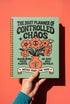A sage green hardcover spiral planner for 2027-2028 with the title "The 2027 Planner of Controlled Chaos" in a funky, retro 70s-style font. The cover features a groovy illustration of orange and pink flowers in a vase, with one flower depicted as an eyeball. The text reads "Bloom where you're planted. Or just wilt artfully." and "Notes from the Void." The planner has a gold metal wire-o binding. The back cover shows a decorative floral frame with a "Property Of" section