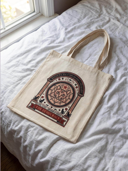 A beige canvas tote bag featuring a vintage-style circular crest design in rust orange and black. The text reads "Society of Childless Cat Dads" in an arch over a central illustration of a tabby cat face inside a geometric star and hexagon symbol. Below the cat, the text reads "Loyal to Cats, Suspicious of Fascists." A banner at the bottom reads "For Democracy." The design features occult and whimsigoth elements like crescent moons, columns, and fish bones.