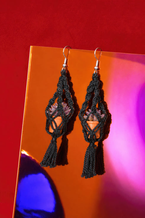 A pair of black tassel earrings with a potted plant design, displayed against a vibrant red background.