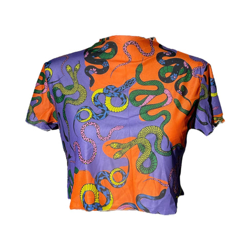 A vibrant see-through mesh crop top with a high mock neck and short sleeves. The shirt features a bold, all-over snake print pattern with colorful green, blue, yellow, and black serpents on a striking orange and purple color-block background. This Y2K-style top is a statement piece of witchy, goth, and alternative clothing, perfect for a maximalist rave outfit or edgy festival fashion from a black owned shop.