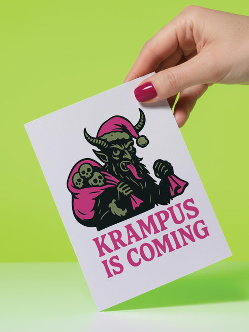 A 5x7 inch Krampus Christmas card shown in two paper finishes. The front features a stylized illustration of the folklore demon Krampus with green skin, black horns, and a pink Santa hat. He carries a large pink sack filled with three skulls over his shoulder. Below him, the words "KRAMPUS IS COMING" are printed in bold pink text. The inside of the card is blank on the left and has the printed text "Hope you've been beautifully bad this year." on the right.
