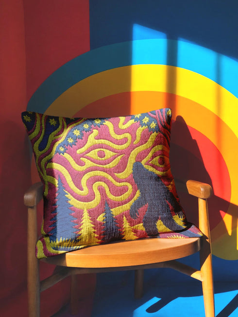 A square decorative throw pillow cover featuring a trippy retro Bigfoot scene. A large navy blue silhouette of Sasquatch walks through a forest of navy and yellow pine trees. The background is a psychedelic swirling sky in vibrant pink and yellow, featuring a large stylized eye and a cluster of small yellow stars. The flat two dimensional print visually mimics the textured look of heavy needlepoint embroidery. The pillow features a sleek hidden zipper on the bottom. No insert is included.