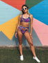 Psychedelic bikini bottom with multicolored tiger print on a neon leopard background; mid-rise cut with black lining and vibrant color blocking in purple, blue, pink, orange, and cream; made of recycled polyester and spandex blend with cheeky fit.
