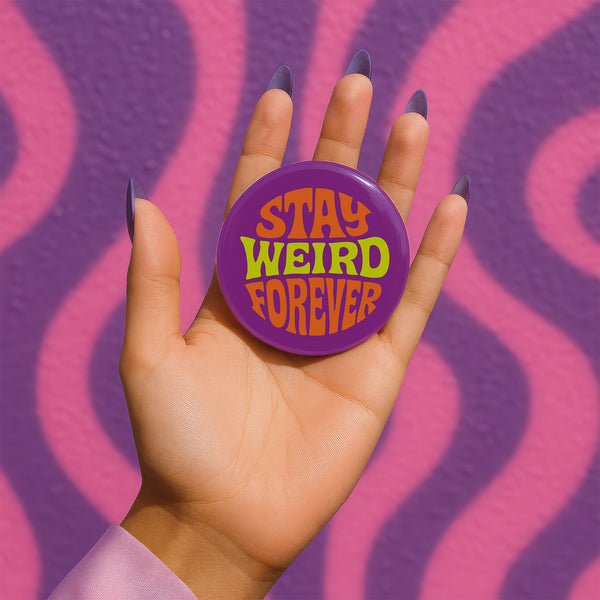 A round metal pin button with a glossy finish and a vibrant purple background. The phrase 'STAY WEIRD FOREVER' is written across the center in a wavy, psychedelic 70s-style font. The words 'STAY' and 'FOREVER' are a bold, bright orange, while the word 'WEIRD' is rendered in a contrasting, electric chartreuse lime green. The button has a smooth, scratch-resistant mylar surface covering the design. The pin is shown in its 2-inch and 3-inch variations, highlighting its circular shape and durable construction.