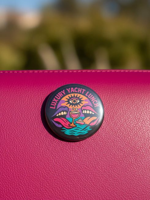 A round 1.75 inch glossy pinback button featuring satirical psychedelic artwork on a black background. The text LUXURY YACHT LUNCH arches over the top in bold neon pink letters. The central design depicts a surreal scene with a stylized third-eye sun, large open mouths with tongues sticking out, a smiling cocktail glass character, and a small white yacht floating on wavy teal water. The color palette includes retro 70s neon pink, orange, purple, and teal. The button has a safety pin backing.