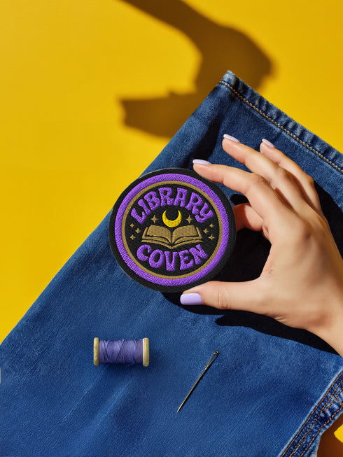 3-inch round embroidered iron-on patch in a cotton–polyester blend; black base with purple ring and gold accent ring; open book, gold crescent moon, tiny stars; curved purple text reads “LIBRARY COVEN”; merrowed edge. Witchy book lover patch for librarians, dark academia, and BookTok covens; makes a great gift for readers, witches, and spooky library pals.