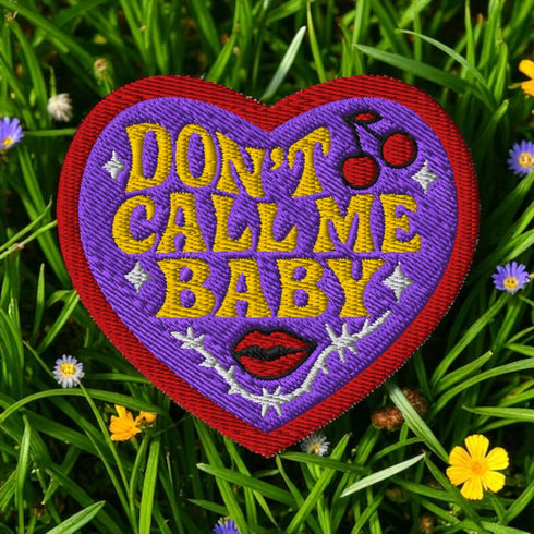 A heart-shaped embroidered patch with a thick red border and a vibrant purple background. In the center, the phrase "DON’T CALL ME BABY" is stitched in a bold, golden-yellow block font. The patch is decorated with small white sparkles, a pair of red cherries with a black musical note, a pair of red lips, and a grey thorny vine design below the text. The overall design has a rebellious, retro, punk aesthetic with a handmade, textured look.