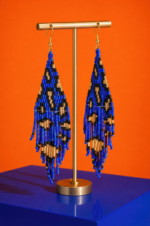 A pair of blue and gold beaded earrings with long, dangling designs, displayed on a gold stand against an orange background.