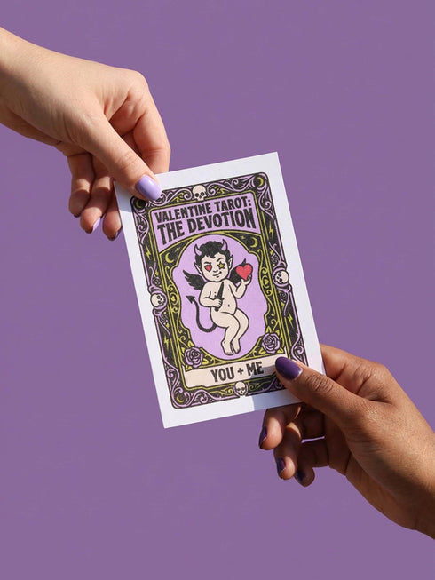 A vertical folded greeting card featuring a pastel goth tarot design titled The Devotion. The central illustration shows a pale devil imp with black wings and horns, holding a red heart in the left hand and a black dagger in the right. The imp has one star eye and one heart eye. The background is lavender purple. A black border frames the scene with teal vines, skulls, gold crescent moons, and roses. The bottom banner reads YOU + ME. The finish is shimmering pearlescent.