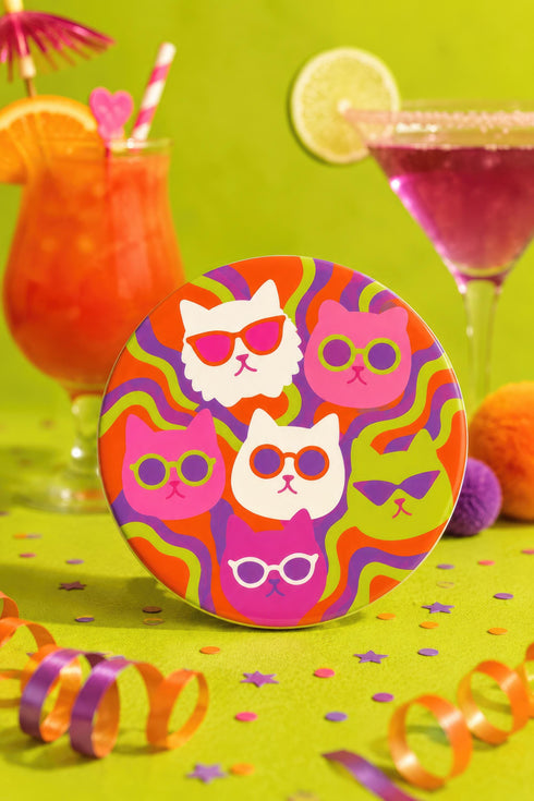A close-up photograph of a glossy round ceramic coaster featuring a retro 70s psychedelic design. The artwork displays a cluster of six stylized cat face illustrations, each wearing a different pair of sunglasses including retro round and cat-eye shapes. The cats are colored in solid shades of white, hot pink, purple, and lime green. They are set against a vibrant background of wavy, swirling lines in bright orange, red, and purple. The top surface has a highly reflective clear glaze.