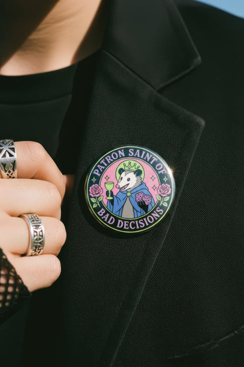 A round glossy metal pinback button featuring a flat 2D print illustration of an opossum wearing a blue cloak and a green crown. The opossum holds a neon green goblet in one paw and two purple dice in the other. A bright pink circle sits behind the opossum, surrounded by a black outer ring. Purple text reads PATRON SAINT OF BAD DECISIONS curved around the edge. Two pink roses with green leaves are on the sides. The design uses bold black outlines and bright neon colors.