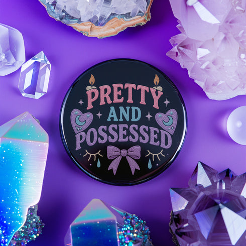 Glossy black pinback button featuring pastel-style text that reads “Pretty and Possessed” with a bow, candles, hearts, and sparkles.