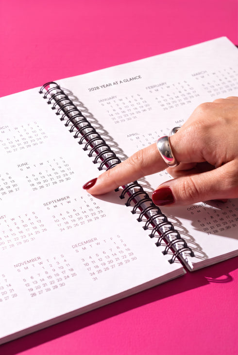 A hand wearing a ring is holding a spiral-bound notebook with a calendar page visible, against a pink background.