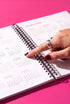 A hand wearing a ring is holding a spiral-bound notebook with a calendar page visible, against a pink background.