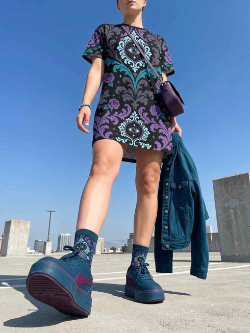 Cotton t-shirt dressA short-sleeve structured t-shirt dress with a black background. The dress features a bold, vertically stacked repeating graphic print. The design includes intricate damask patterns in teal and grey, purple roses with teal leaves, and a central evil eye motif set inside an ornate frame with small four-point stars. The heavyweight fabric holds a clean, straight cut above the knee with zero drape, maintaining a sculptural, slightly boxy silhouette.
