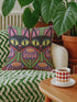 A square decorative pillow cover featuring a vibrant, symmetrical flat printed design that mimics the look of textured embroidery. Two black cats with large yellow eyes and pink noses face forward against a bright red background. Below the cats are large purple and pink flowers with green stems. Above the cats are green cactus shapes. The artwork uses bold, colorful outlines to create an optical illusion of stitching, though the fabric itself is smooth.