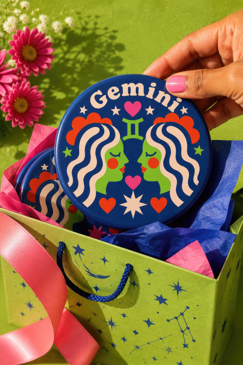 A close-up photograph of a round, glossy ceramic coaster featuring a bold retro pop art design. The background is a deep royal blue. In the center are two symmetrical profile faces of twins looking at each other, featuring bright green skin, closed eyes, and red cheeks. They have wavy white and pale pink hair with red accents. The word Gemini is arched at the top in a retro white font. The design is decorated with a green Gemini symbol, pink and red hearts, and white starbursts.