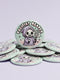 Round pinback button featuring a pastel mint green background with a cartoon grim reaper character in a lavender hood holding a protest sign. The design includes bold black outlined lettering reading “Pacifist Reaper” above the figure and small sparkle accents surrounding it. The finish is glossy and smooth, with a printed illustration style on a metal pin badge surface.
