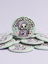 Round pinback button featuring a pastel mint green background with a cartoon grim reaper character in a lavender hood holding a protest sign. The design includes bold black outlined lettering reading “Pacifist Reaper” above the figure and small sparkle accents surrounding it. The finish is glossy and smooth, with a printed illustration style on a metal pin badge surface.