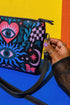 Small vegan leather crossbody purse with a real pebble grain texture and adjustable black strap. The flat printed artwork features a central blue sunburst evil eye motif below a coral pink sacred heart eye, surrounded by pink crescent moons, multicolored stars, flowing teal botanical vines, and wavy lavender and black checkerboard distortion banners. Set against a dark background with silver metal hardware including a top zipper, zipper pull, and strap clips. Measures 8.5 by 7 by 2 inches.