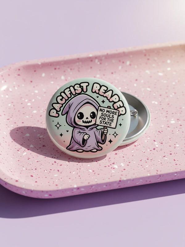Round pinback button featuring a pastel mint green background with a cartoon grim reaper character in a lavender hood holding a protest sign. The design includes bold black outlined lettering reading “Pacifist Reaper” above the figure and small sparkle accents surrounding it. The finish is glossy and smooth, with a printed illustration style on a metal pin badge surface.