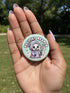 Round pinback button featuring a pastel mint green background with a cartoon grim reaper character in a lavender hood holding a protest sign. The design includes bold black outlined lettering reading “Pacifist Reaper” above the figure and small sparkle accents surrounding it. The finish is glossy and smooth, with a printed illustration style on a metal pin badge surface.