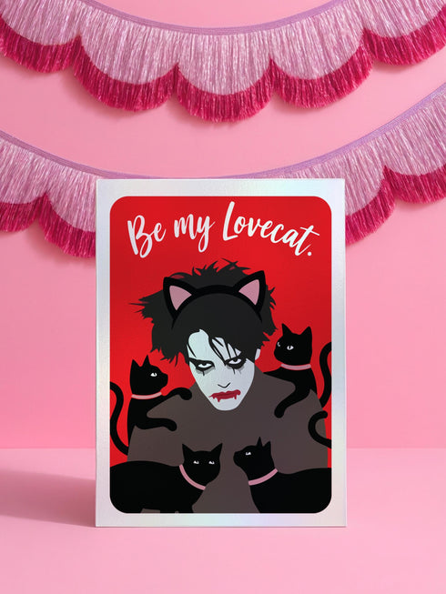 A vertical greeting card featuring a flat vector illustration of Robert Smith from The Cure against a vibrant red background. He has messy black hair, pale skin with smeared lipstick, and wears black cat ears. He is surrounded by four black cats with pink collars. White script text at the top reads "Be my Lovecat." The card is printed on shimmery pearlescent cardstock that catches the light. A high-quality alternative Valentine or anniversary card for goth music lovers and post-punk fans.