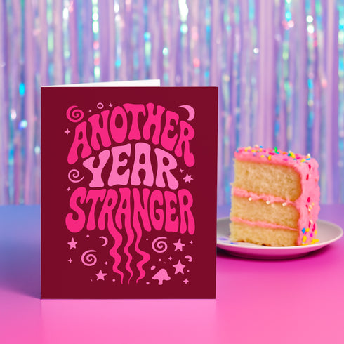A red greeting card with the text "Another Year Stranger" in a whimsical font, placed next to a slice of cake on a plate against a purple background with a shimmering effect.