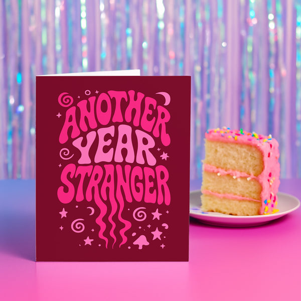A red greeting card with the text "Another Year Stranger" in a whimsical font, placed next to a slice of cake on a plate against a purple background with a shimmering effect.
