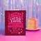 A red greeting card with the text "Another Year Stranger" in a whimsical font, placed next to a slice of cake on a plate against a purple background with a shimmering effect.