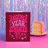A red greeting card with the text "Another Year Stranger" in a whimsical font, placed next to a slice of cake on a plate against a purple background with a shimmering effect.