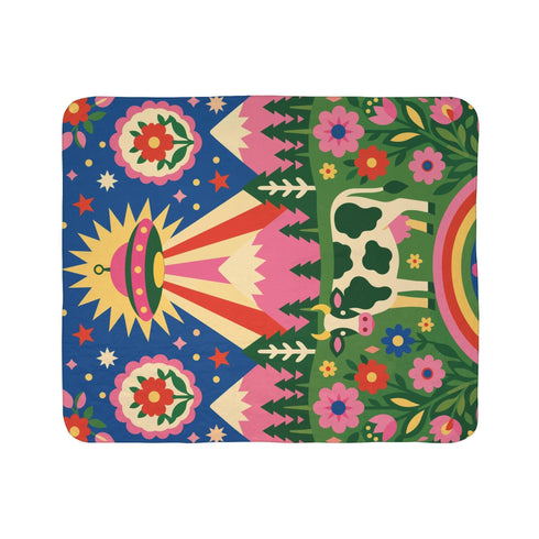 A rectangular velveteen minky blanket with a bright, maximalist design. The top half features a vibrant blue sky with a pink, green, and yellow UFO beaming rays down over hot pink snow-capped mountains. The bottom half shows a grassy green field with a white cow covered in dark spots, surrounded by large, colorful red, pink, and blue retro flowers, pine trees, and a rainbow arch at the very bottom. The reverse side is a fluffy off-white sherpa.