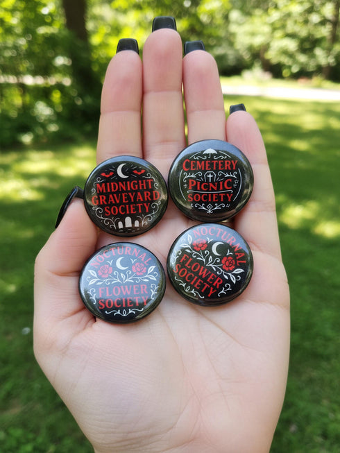 A collection of four glossy black circular pinback buttons featuring striking red and white gothic illustrations. One button shows "Midnight Graveyard Society" text with bats and tombstones. Another reads "Cemetery Picnic Society" with a coffin and umbrella icon. A third features a detailed white skull and red roses with "Memento Mori" text below. The final pin displays "Nocturnal Flower Society" surrounded by floral vines.