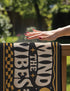 A rectangular floor mat with a dark charcoal black background. The center features the phrase 'MIND THE VIBES' in stylized, wavy cream and golden-mustard retro typography. The text is surrounded by symmetrical motifs: a golden evil eye with sun rays on the top left, a golden crescent moon with a four-point star on the top right, and two cream daisy flowers with golden stems at the bottom corners. The bottom edge has a wavy golden checkerboard pattern. A faux-stitch border frames the art.