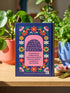 Royal blue hardcover journal featuring a retro folk art design. A center pink arch frames the word VIRGO in golden groovy rounded lettering. Below sits an illustrated maiden with brown skin and dark hair, wearing a pink dress and holding a bundle of golden wheat. A dense floral border wraps the cover with daisies and blooms in orange, marigold yellow, cream white, and pink, accented by green leaves and stems. Matte finish cover with a blue spine reading Virgo. Lined pages inside.