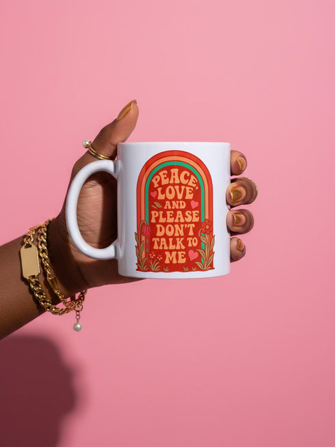 A glossy white porcelain ceramic mug featuring a vibrant maximalist design centered on the exterior. The artwork displays a retro 1970s-inspired rainbow arch composed of concentric bands in burnt orange, mustard yellow, and teal green. Inside the arch, stylized vintage bubble-serif typography in reddish-orange reads PEACE LOVE AND PLEASE DON'T TALK TO ME. The design evokes a groovy, hippie aesthetic mixed with sassy introvert humor.