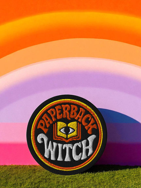 A round, black embroidered patch with a thick, textured merrow border stitched in alternating orange and yellow thread. In the center, the words "PAPERBACK WITCH" are embroidered in a retro 70s-style font. The word "PAPERBACK" is arched across the top in bold orange thread with a thin yellow outline. The word "WITCH" is across the bottom in puffy, bright white thread with a thin yellow outline. Between the words is a central icon of an open book, containing a stylized all-seeing eye.