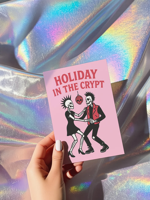 A 5x7 inch greeting card with a vibrant pink background and red retro text reading 'HOLIDAY IN THE CRYPT'. The illustration shows a male and female punk rock skeleton dancing under a red skull disco ball. The female skeleton has a mohawk and wears a black dress. The male skeleton has a mohawk and wears a black jacket with a red vest. Inside, the message reads, 'From our crypt to yours. Eat, drink, and be scary.' The style is a graphic, punk aesthetic.
