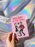 A 5x7 inch greeting card with a vibrant pink background and red retro text reading 'HOLIDAY IN THE CRYPT'. The illustration shows a male and female punk rock skeleton dancing under a red skull disco ball. The female skeleton has a mohawk and wears a black dress. The male skeleton has a mohawk and wears a black jacket with a red vest. Inside, the message reads, 'From our crypt to yours. Eat, drink, and be scary.' The style is a graphic, punk aesthetic.