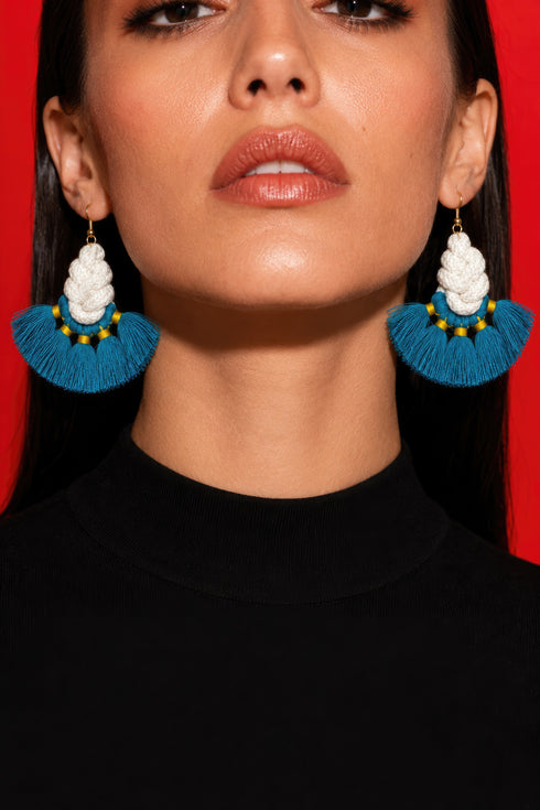 A woman wearing large, blue earrings with white tassels against a red background.