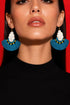 A woman wearing large, blue earrings with white tassels against a red background.