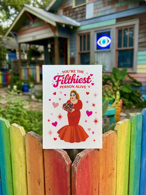 A 5x7 inch pearlescent silver greeting card featuring a flat vector illustration of the drag queen Divine in a red mermaid gown holding a heart-shaped box of chocolates. The text above reads "You're The Filthiest Person Alive" in bold red script with pink sparkles. The card has a shimmering finish that reflects light. Inside text reads "...and I love it." The back features the Honey Dazed logo. Includes a matching envelope.