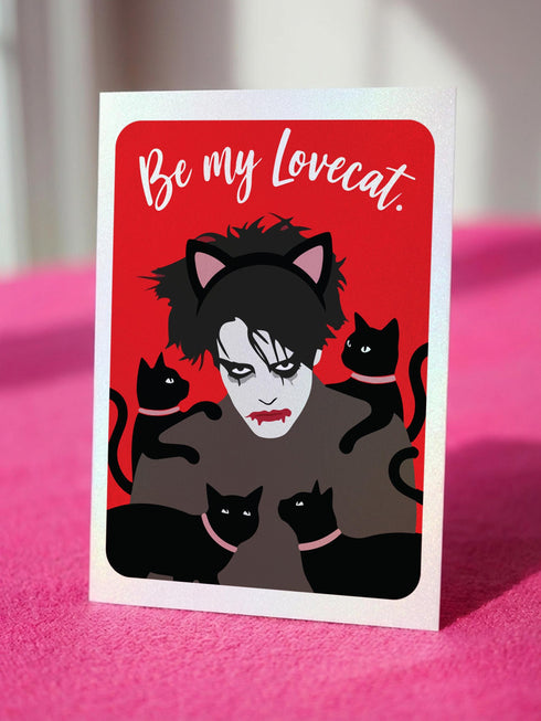 A vertical greeting card featuring a flat vector illustration of Robert Smith from The Cure against a vibrant red background. He has messy black hair, pale skin with smeared lipstick, and wears black cat ears. He is surrounded by four black cats with pink collars. White script text at the top reads "Be my Lovecat." The card is printed on shimmery pearlescent cardstock that catches the light. A high-quality alternative Valentine or anniversary card for goth music lovers and post-punk fans.