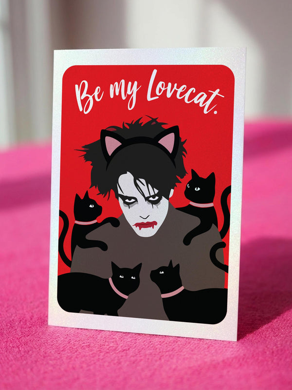 A vertical greeting card featuring a flat vector illustration of Robert Smith from The Cure against a vibrant red background. He has messy black hair, pale skin with smeared lipstick, and wears black cat ears. He is surrounded by four black cats with pink collars. White script text at the top reads "Be my Lovecat." The card is printed on shimmery pearlescent cardstock that catches the light. A high-quality alternative Valentine or anniversary card for goth music lovers and post-punk fans.