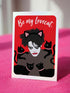 A vertical greeting card featuring a flat vector illustration of Robert Smith from The Cure against a vibrant red background. He has messy black hair, pale skin with smeared lipstick, and wears black cat ears. He is surrounded by four black cats with pink collars. White script text at the top reads "Be my Lovecat." The card is printed on shimmery pearlescent cardstock that catches the light. A high-quality alternative Valentine or anniversary card for goth music lovers and post-punk fans.