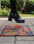 A rectangular doormat featuring a vibrant 2D flat print designed to look like textured yarn embroidery. The central graphic showcases a bright green alien with large black eyes, wearing a pink neck bandana and a highly decorated red, pink, and blue sombrero cowboy hat. The alien has one green hand raised in a friendly wave. The background is a psychedelic pattern of wavy, flowing rainbow stripes in yellow, orange, red, pink, blue, and green. The mat has a sleek black outer edge trim.